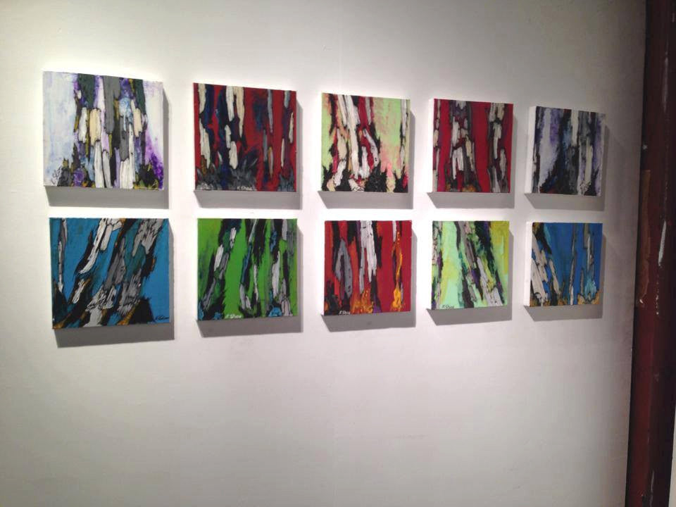 10 colorful original paintings hung on a gallery wall horizontally.