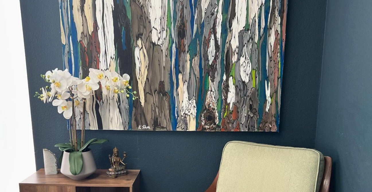An abstract canvas wall art print with vertical textured brushstrokes in blue, white, gray, brown, and green, hanging on a dark blue wall above a wooden side table with a potted white orchid and a light green armchair.