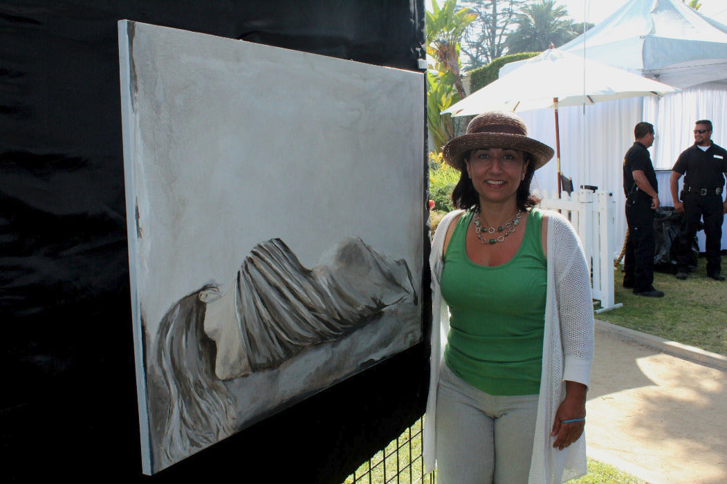 A woman wearing a brown hat, green top, and white cardigan stands next to a large canvas painting depicting a draped figure, set at an outdoor art fair with tents and people in the background.